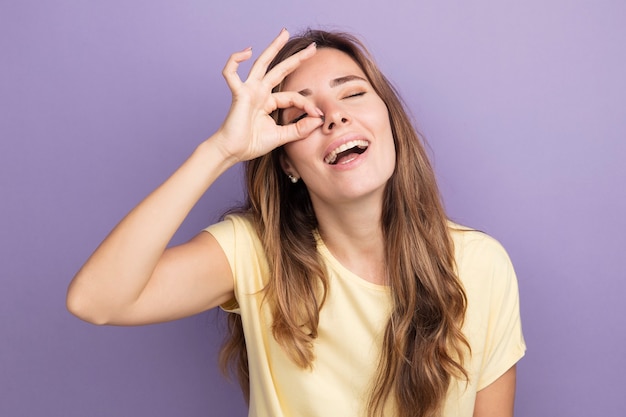 Gelukkige jonge mooie vrouw in beige t-shirt die ok teken doet die door dit teken kijkt