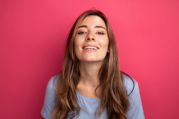 Gelukkige jonge mooie vrouw in blauw t-shirt die naar de camera kijkt met een glimlach op het gezicht dat over roze staat