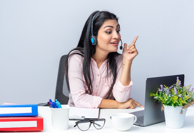 Gelukkige jonge mooie vrouw in casual kleding met koptelefoon en microfoon zittend aan de tafel met laptop met videogesprek over witte achtergrond werken op kantoor