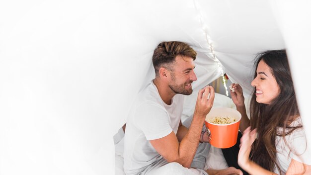 Gratis foto gelukkige jonge paarzitting binnen het gordijn op bed die popcorn eten