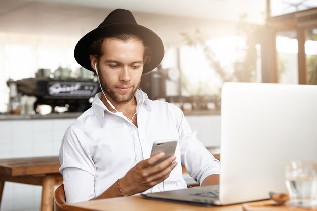 Gelukkige jonge student modieuze hoed dragen en oortelefoons die aan muziek op zijn mobiele telefoon luisteren, terwijl ontspannend binnen bij gezellige cafetaria, die bij lijst met laptop computer zitten.