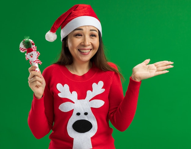 Gelukkige jonge vrouw die de hoed van de Kerstmissanta en rode sweater draagt die het riet van het Kerstmissuikergoed opzij kijkt met glimlach op gezicht die zich over groene achtergrond bevinden