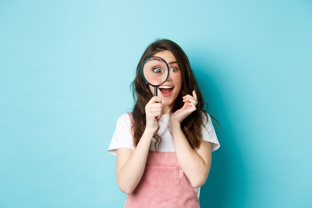 Gelukkige jonge vrouw die door vergrootglas kijkt met een opgewonden gezicht, iets gevonden of zoekt, staande over een blauwe achtergrond