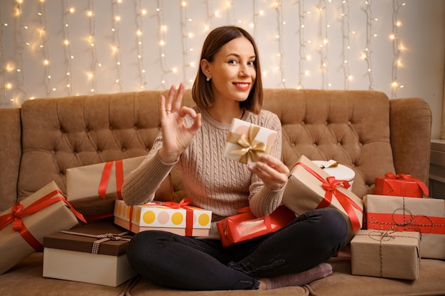 Gelukkige jonge vrouw die een cadeautje houdt dat ok teken toont dat door giftdozen wordt omringd die gekruiste benen zitten