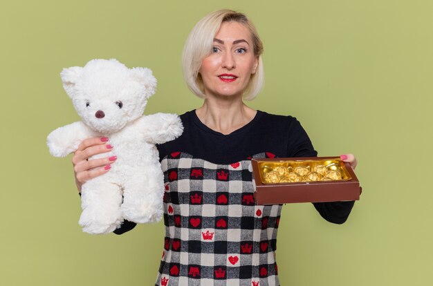 Gelukkige jonge vrouw die in mooie kleding teddybeer en chocoladesuikergoed houdt als giften glimlachend vrolijk viert de dag van de internationale vrouw die zich over groene muur bevindt