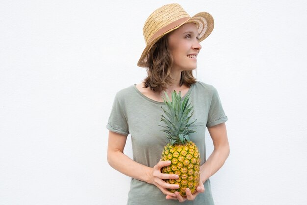 Gelukkige jonge vrouw die in strohoed gehele ananas houdt