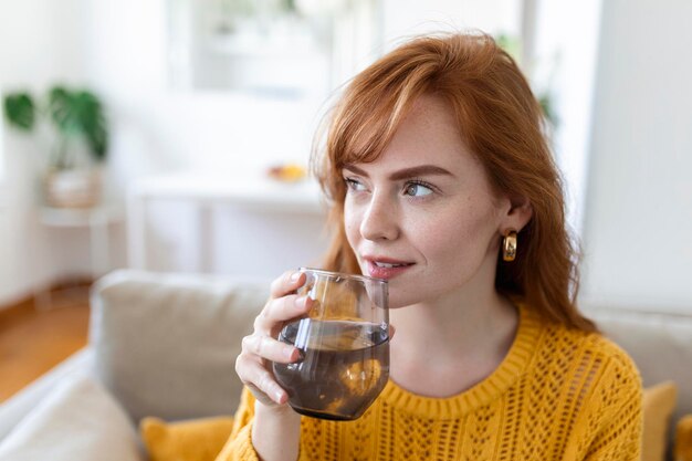 Gelukkige jonge vrouw die lacht terwijl ze thuis een glas water vasthoudt Vrouw op de bank in de woonkamer Ontspannen en glimlachend terwijl ze een glas water drinkt Gezondheidsvoordelen van voldoende water drinken