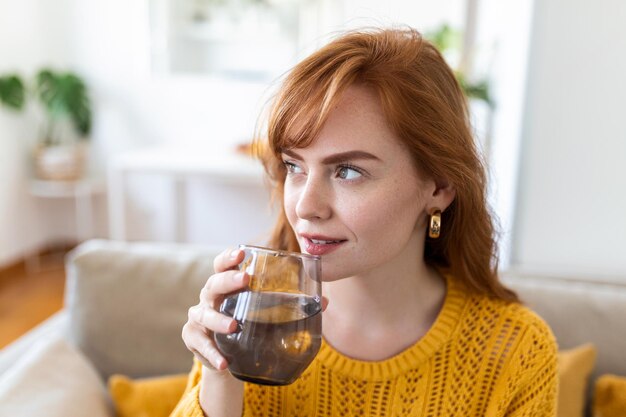 Gelukkige jonge vrouw die lacht terwijl ze thuis een glas water vasthoudt Vrouw op de bank in de woonkamer Ontspannen en glimlachend terwijl ze een glas water drinkt Gezondheidsvoordelen van voldoende water drinken