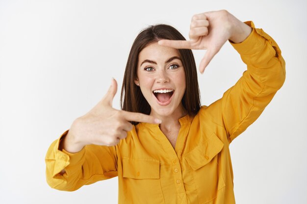 Gelukkige jonge vrouw die moment vastlegt, cameragebaar maakt en glimlacht, staande over een witte muur