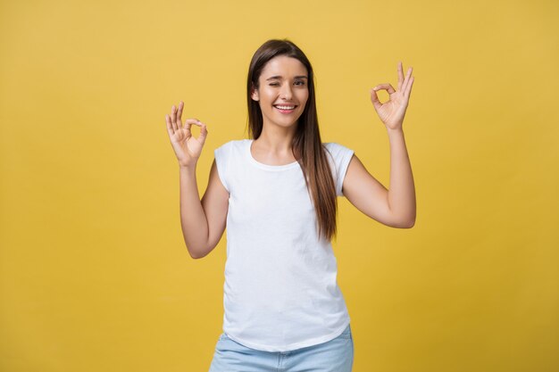 Gelukkige jonge vrouw die ok teken met vingers toont die geïsoleerd op een gele achtergrond knipogen