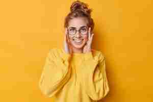 Gratis foto gelukkige jonge vrouw houdt handen op gezicht glimlacht breed toont witte tanden draagt ronde bril voor zicht casual jumper geïsoleerd over gele achtergrond. menselijk gezichtsuitdrukkingen en emoties concept
