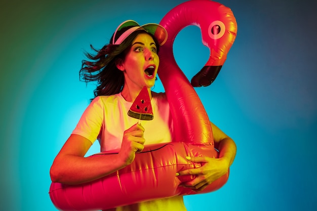 Gelukkige jonge vrouw in een pet en in een rubberen flamingo met het snoepje op trendy blauwe neon