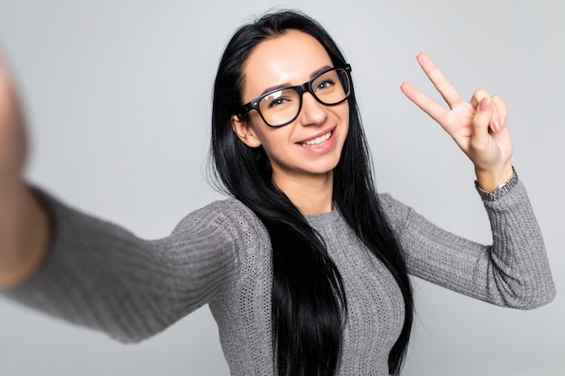 Gelukkige jonge vrouw in glazen met stralende glimlach maken selfie met v-teken geïsoleerd op grijze muur