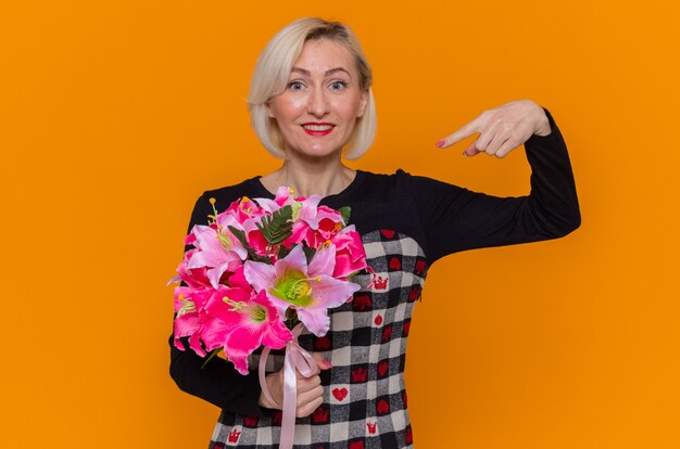 Gelukkige jonge vrouw in mooie jurk met boeket bloemen wijzend met wijsvinger naar het glimlachend vrolijk internationale Vrouwendag vieren staande over oranje muur
