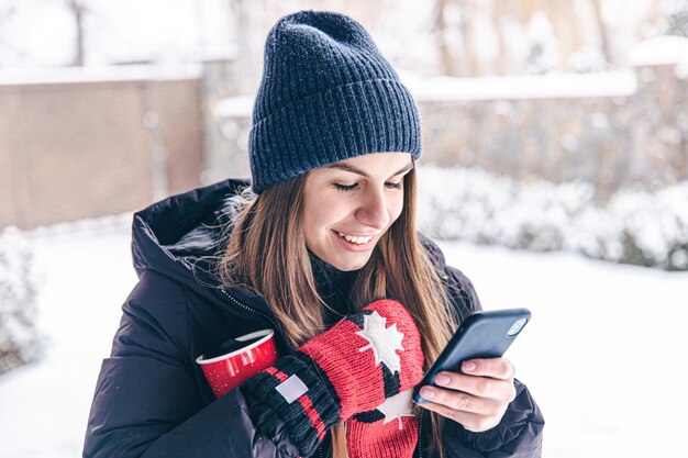 Gelukkige jonge vrouw in wanten met een kopje kijkt naar het telefoonscherm