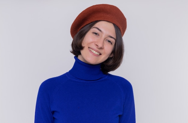 Gelukkige jonge vrouw met kort haar in blauwe coltrui die baret draagt die voorzijde glimlachend vrolijk status over witte muur bekijkt