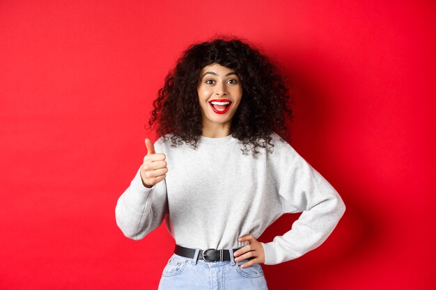 Gelukkige jonge vrouw met krullend haar die goed werk prees, goed gedaan zegt en duim omhoog gebaar toont, keurt en prijst, staande op rode achtergrond.