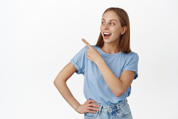 Gelukkige jonge vrouw, student wijzend en kijkend naar de linkerbovenhoek met zorgeloos gezicht, lachend om grappige advertentie, verkoopbanner, staande over witte muur