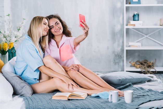 Gelukkige jonge vrouwelijke vrienden die op bed zitten die selfie met smartphone nemen