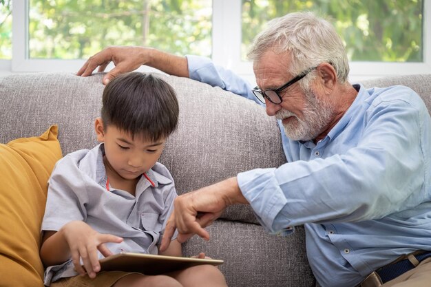 Gelukkige jongen kleinzoon speelspel op tablet met oude senior man grootvader thuis