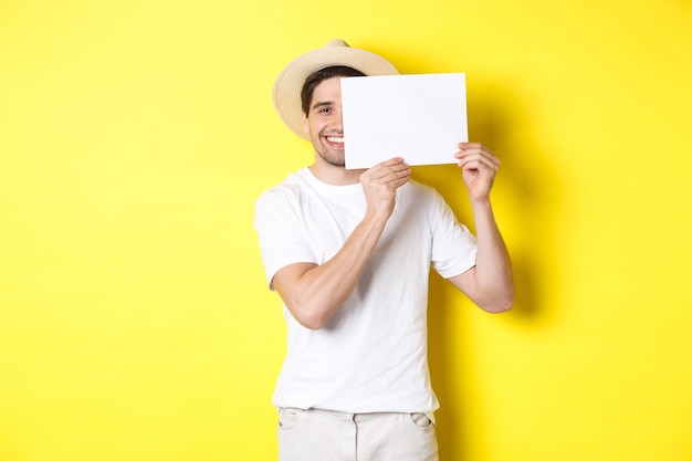 Gelukkige kerel op vakantie die leeg stuk papier voor uw embleem toont, teken dichtbij gezicht houdt en glimlacht, staand tegen gele achtergrond.