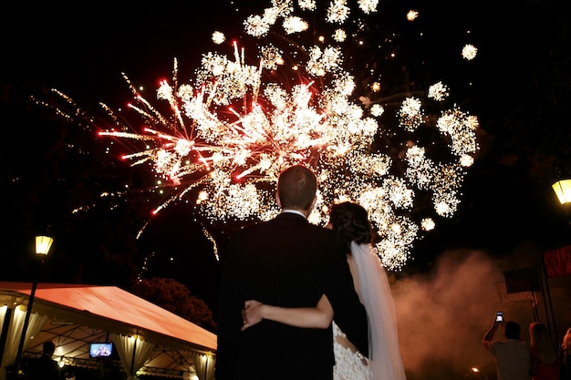 Gelukkige knuffelbruid en bruidegom kijken naar mooie kleurrijke vuurwerk nachtelijke hemel