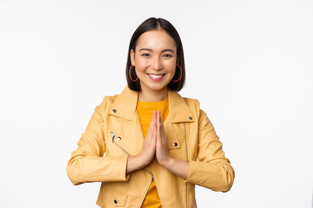 Gelukkige Koreaanse vrouw die er hoopvol uitziet en om hulp vraagt, smekend staat met namaste-gebaar en glimlacht terwijl ze op een witte achtergrond staat