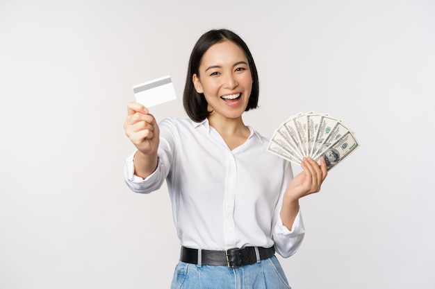 Gelukkige Koreaanse vrouw met creditcard en gelddollars glimlachend en lachend poserend tegen witte studioachtergrond