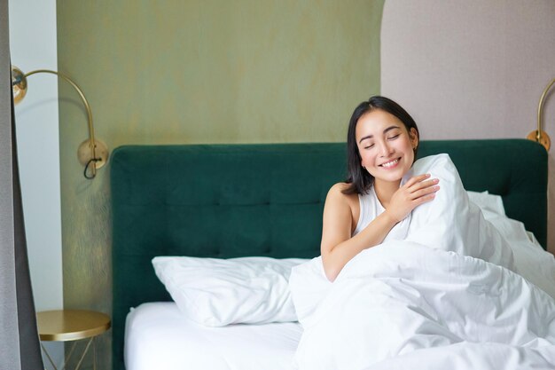 Gelukkige koreaanse vrouw wordt wakker in haar hotelkamer, liggend in bed onder knusse warme dekens, witte lakens enj
