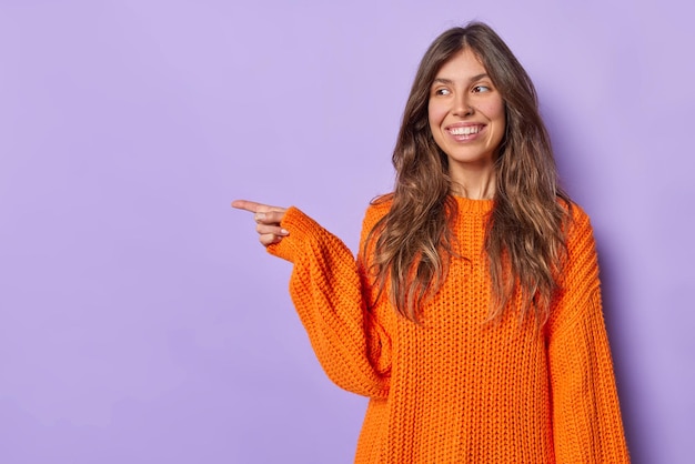 Gelukkige langharige vrouw wijst weg toont richting van iets gekleed in gebreide oranje trui toont plaats voor uw advertentie geïsoleerd over paarse achtergrond. manier om banner te verkopen