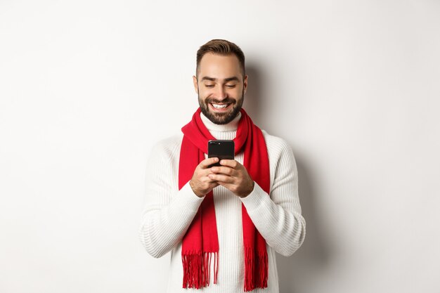 Gelukkige man die lacht tijdens het lezen van bericht op mobiele telefoon, staande in wintertrui en rode sjaal tegen witte achtergrond