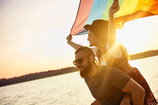 Gelukkige man die vrouw met regenboogvlag over zee draagt