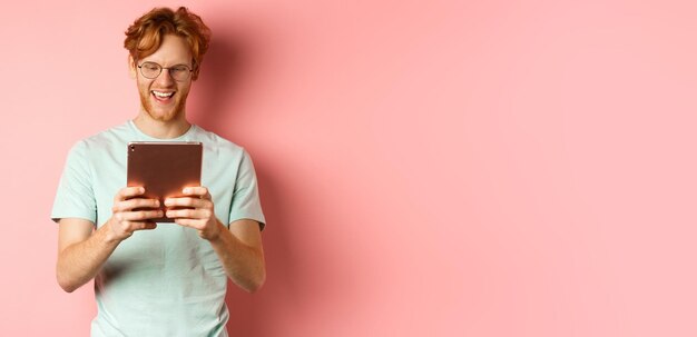 Gratis foto gelukkige man met rood haar en baard die het leesscherm van een digitale tablet gebruikt en glimlacht terwijl hij over roze staat