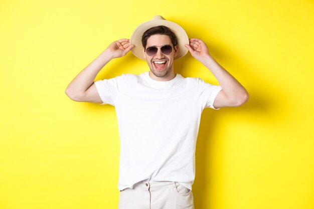 Gelukkige man op vakantie, met strohoed en zonnebril, glimlachend terwijl hij tegen een gele achtergrond staat.