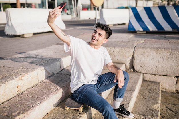 Gelukkige mannelijke skateboarderzitting op skateboard die selfie met mobiele telefoon nemen