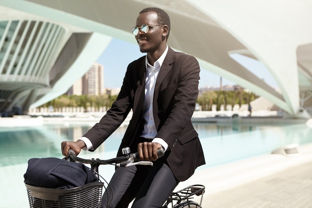 Gelukkige milieubewuste Afro-Amerikaanse werknemer in zwart formeel pak en zonnebril die de fiets kiest boven het openbaar vervoer of de auto om naar kantoor te gaan, genietend van een rit in een stedelijke omgeving