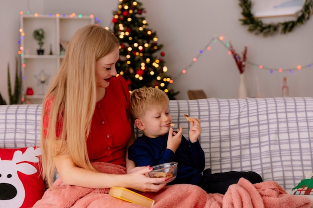 gelukkige moeder in rode jurk met haar kleine kind onder deken kind voeden met koekjes in een ingerichte kamer met kerstboom op de achtergrond