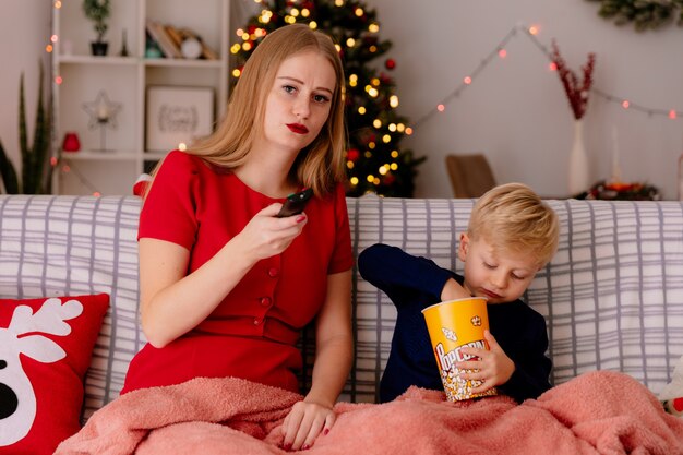 Gelukkige moeder in rode jurk met haar kleine kind zittend op een bank onder deken met emmer popcorn samen tv kijken in een ingerichte kamer met kerstboom in de muur