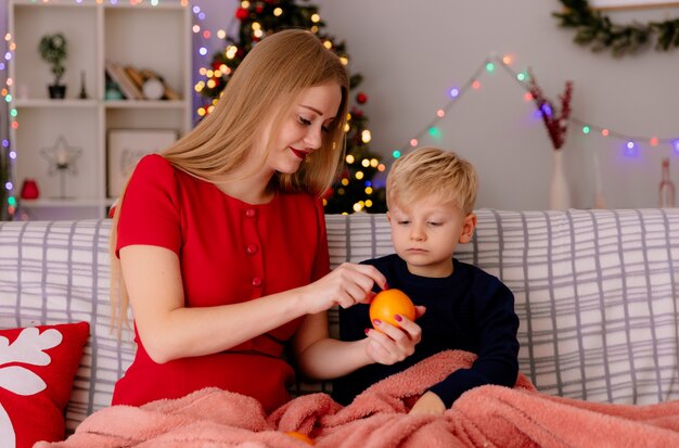 gelukkige moeder in rode jurk oranje geven aan haar kleine kind zittend op een bank onder deken in een ingerichte kamer met kerstboom op de achtergrond