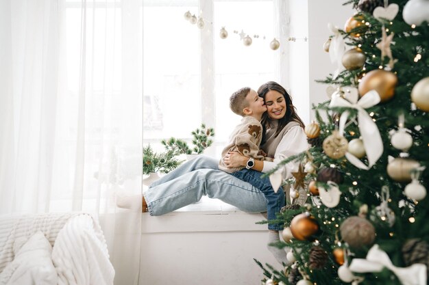 Gelukkige moeder met zoon zit op de vensterbank bij de kerstboom