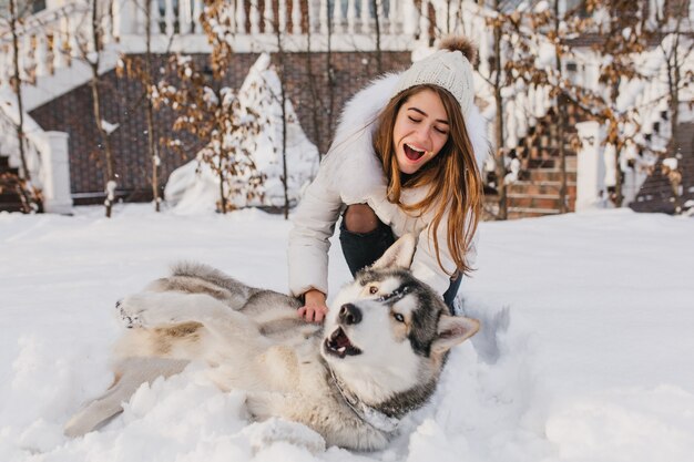 Gelukkige momenten in de winter van geweldige jeige vrouw spelen met husky hond in de sneeuw. Heldere positieve emoties, echte vriendschap, huisdierenliefde, beste vrienden, glimlachen, plezier maken, wintervakanties.
