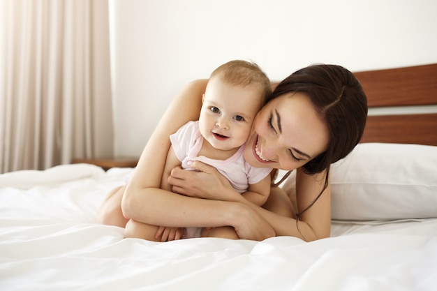 Gelukkige mooie moeder in nachtkleding die op bed met haar babydochter ligt die het glimlachen omhelst.