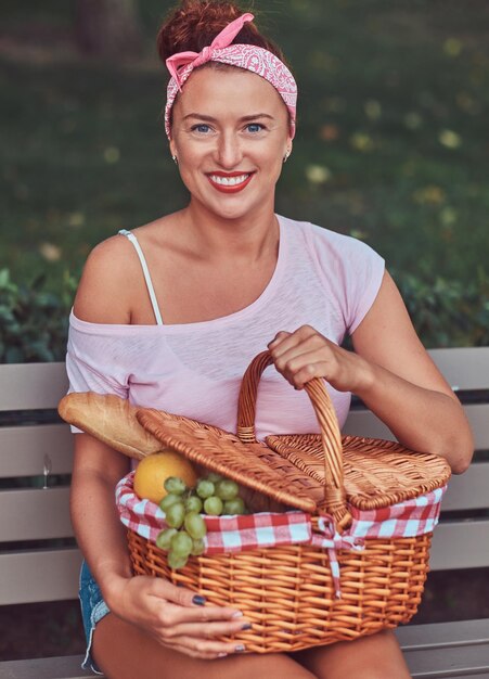 Gelukkige mooie roodharige vrouw die vrijetijdskleding draagt, houdt een picknickmand vast terwijl ze op een bankje in het park zit.