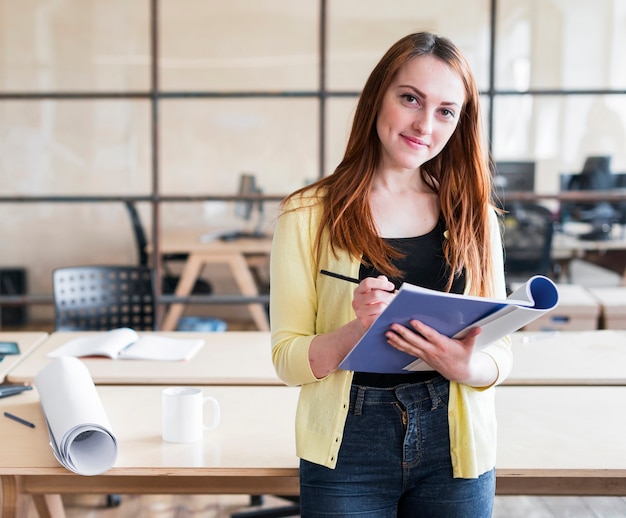 Gelukkige mooie vrouw die op het boek en het potlood van de bureauholding op het werk leunen