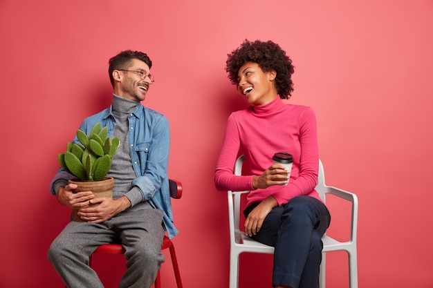 Gelukkige multi-etnische vrouw en man hebben een prettig gesprek, kijken elkaar aan en poseren op stoelen