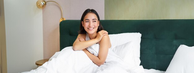 Gratis foto gelukkige ochtenden lachende aziatische vrouw wordt wakker in haar bed, kijkt naar buiten en voelt zich enthousiast