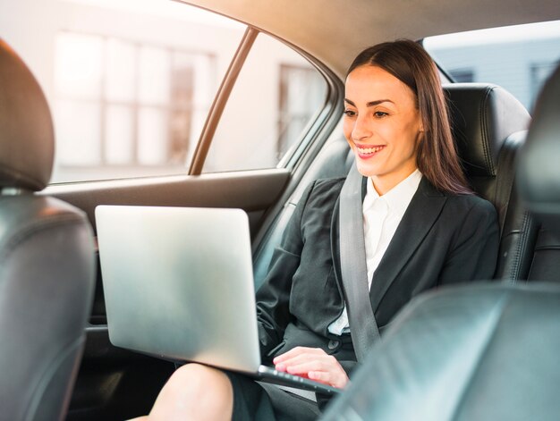 Gelukkige onderneemster die door auto reist die laptop met behulp van