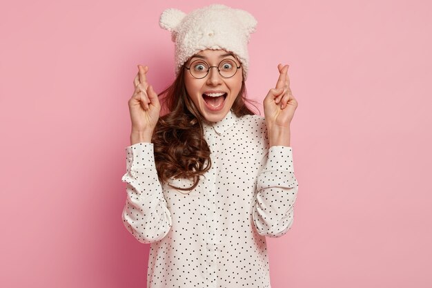 Gelukkige optimistische dame gelooft oprecht in geluk, bidt voor beter, houdt de vingers gekruist, draagt een optische bril, een hoofddeksel en een polkadot-shirt, geïsoleerd over een roze muur. Wens maken