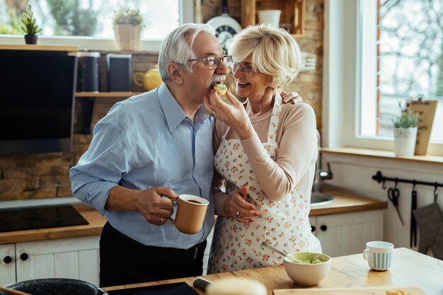 Gelukkige oudere man die zijn vrouw omhelst terwijl ze kookt en hem voedt in de keuken