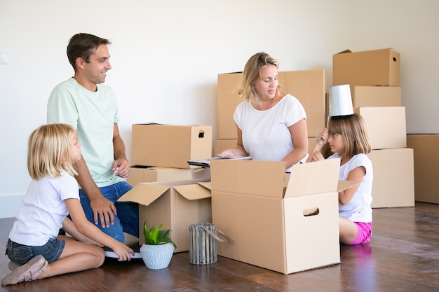 Gelukkige ouders en kleine meisjes die plezier hebben tijdens het uitpakken van dingen in een nieuw appartement, zittend op de vloer en objecten uit open dozen halen